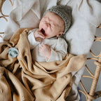 Newborn baby wrapped in a beige blanket with a textured surface and a pacifier from the Frigg 6-pack pacifiers in silicone in Earthy wonder