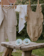 Two baby pacifiers from the Frigg Daisy Bloom pacifier 2-pack in silicone in Little darling cream on a wooden bench with clothes hanging in the background