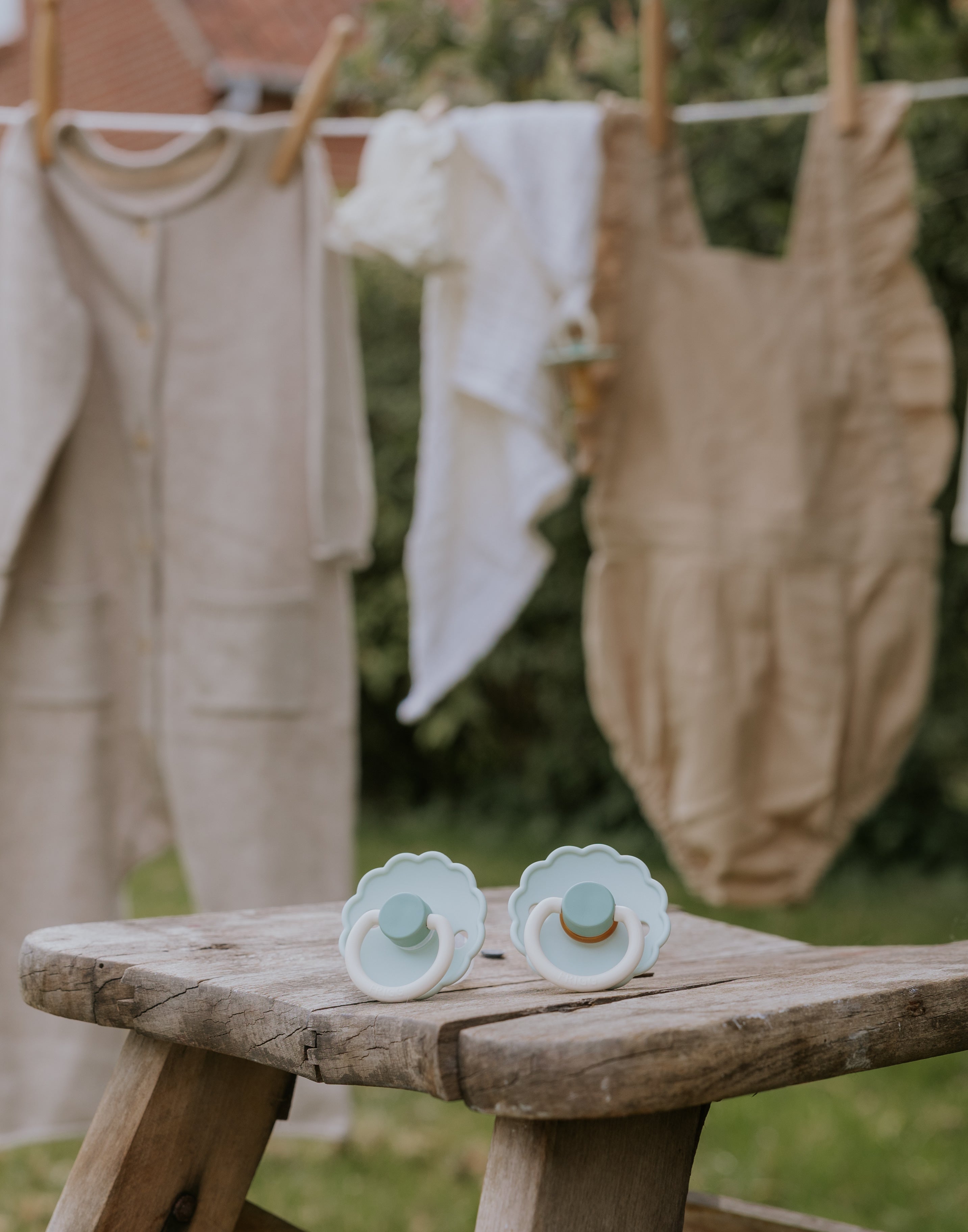 Two baby pacifiers from the Frigg Daisy Bloom pacifier 2-pack in silicone in Little darling cream on a wooden bench with clothes hanging in the background