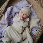 Newborn baby in a beige outfit and hat, holding a pacifier from the Frigg Daisy pacifier 2-pack in silicone in Powder blush pink cream, wrapped in a purple blanket in a woven basket.