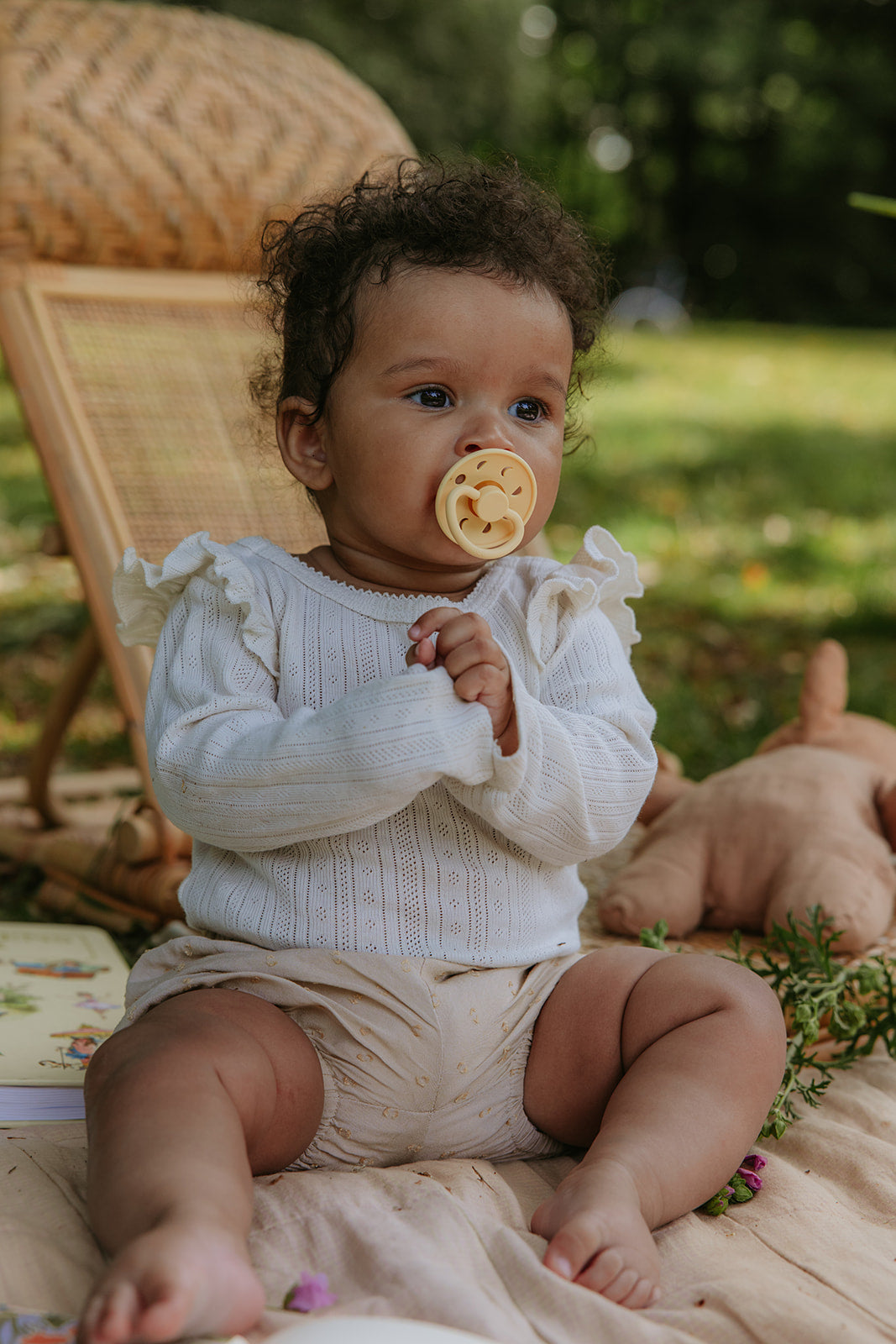 Baby sitting outdoors with a pacifier from the Frigg Moon pacifier 2-pack in silicone in Pale daffodil soft lilac wearing a white sweater and beige shorts.
