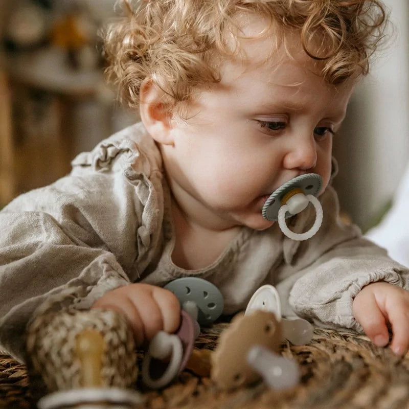 Baby wearing a beige outfit with a pacifier from the Frigg Rope Night pacifier 2-pack in latex in Stone blue sage styled with other Frigg pacifiers