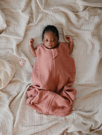 Newborn baby in a pink outfit lying on a textured beige blanket with a  pacifier from the Frigg Rope pacifier 2-pack in silicone in Blush croissant