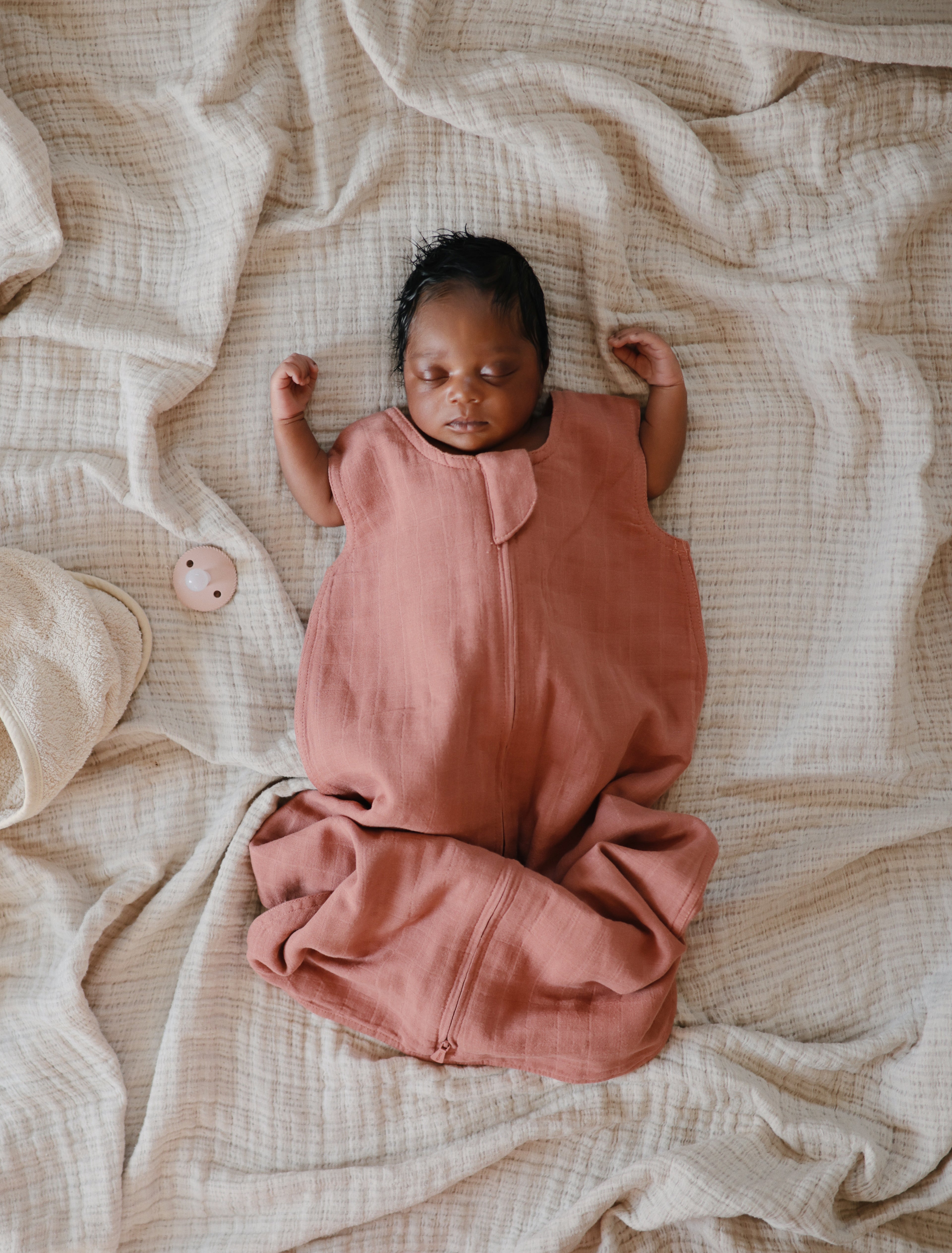 Newborn baby in a pink outfit lying on a textured beige blanket with a  pacifier from the Frigg Rope pacifier 2-pack in silicone in Blush croissant