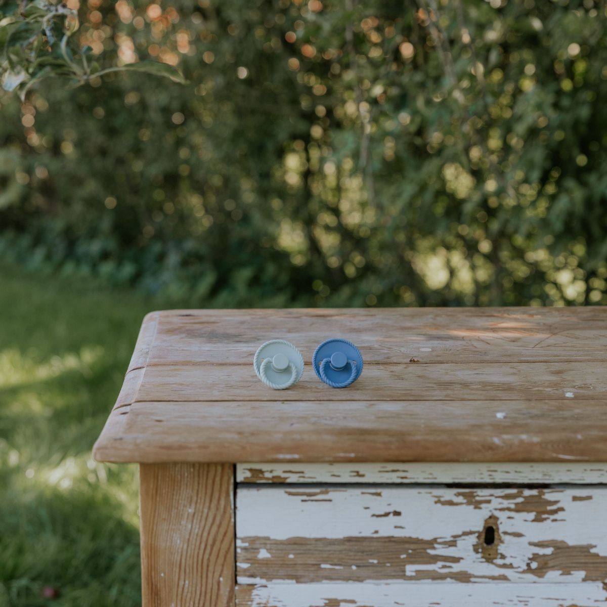 Wooden bench with two pacifiers from the Frigg Rope pacifier T1 2-pack in silicone in Ocean view Sage on top, blurred greenery in the background