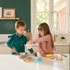 Two children in a kitchen setting playing with Janod's my pastry workshop wooden play set