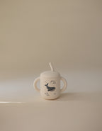 Mushie training cup with straw in Whales print on a beige background