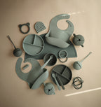 Mushie silicone bib in Cambridge blue styled with green silicone baby utensils and accessories on a beige background