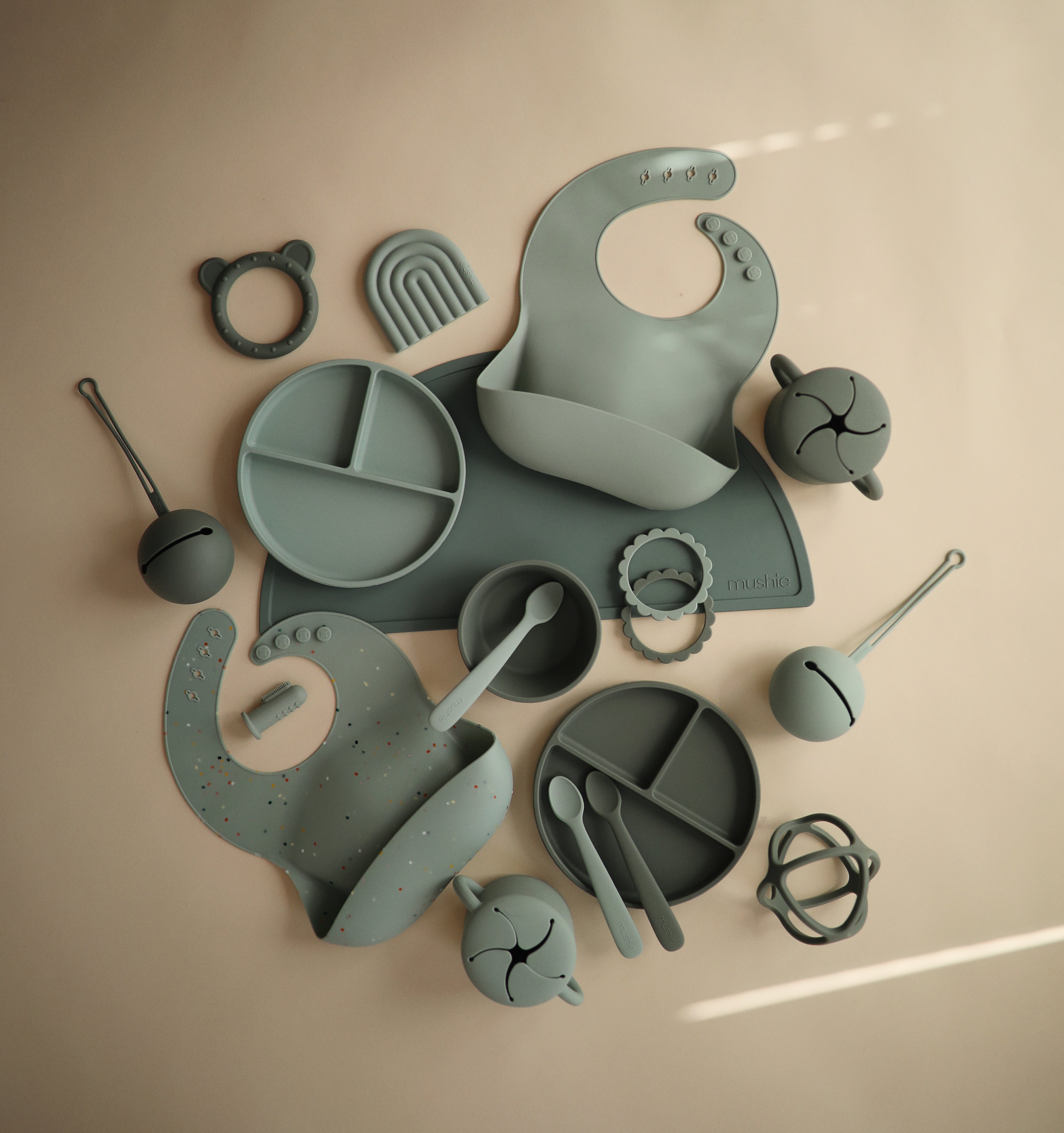 Mushie silicone bib in Cambridge blue styled with green silicone baby utensils and accessories on a beige background