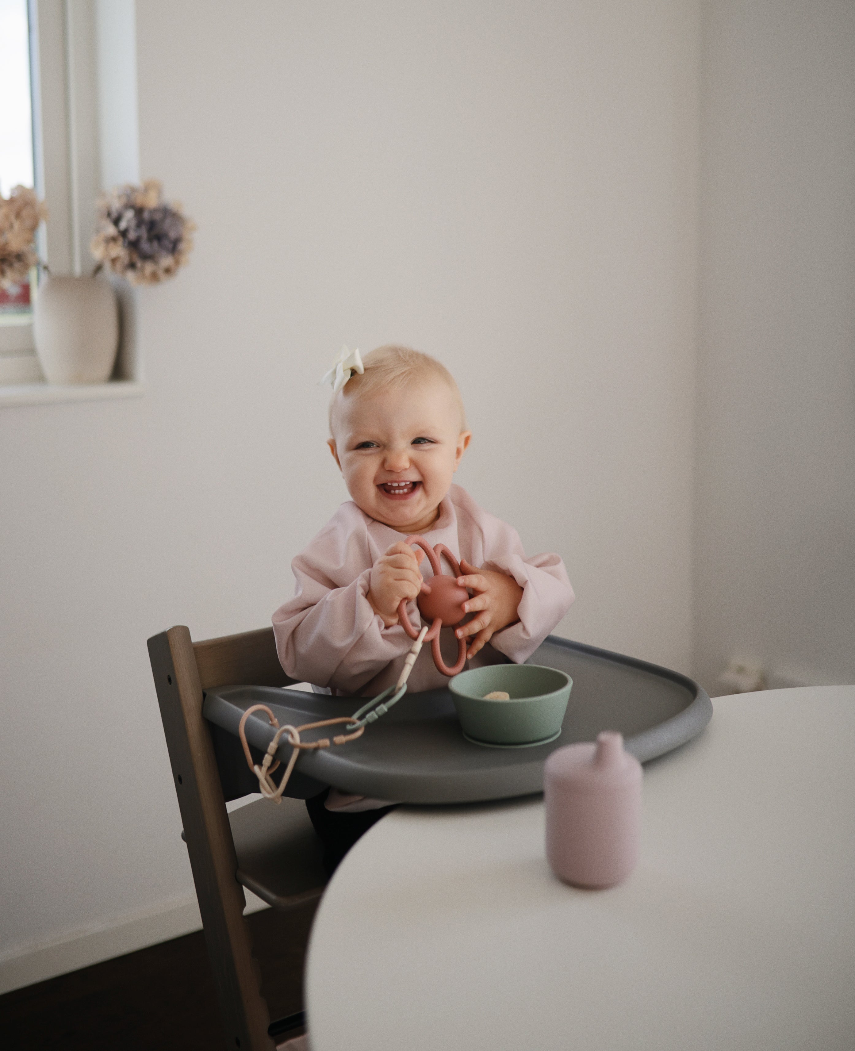 Smiling baby in a high chair wearing a Mushie long sleeve bib in Blush, styled with baby toys and feeding accessories