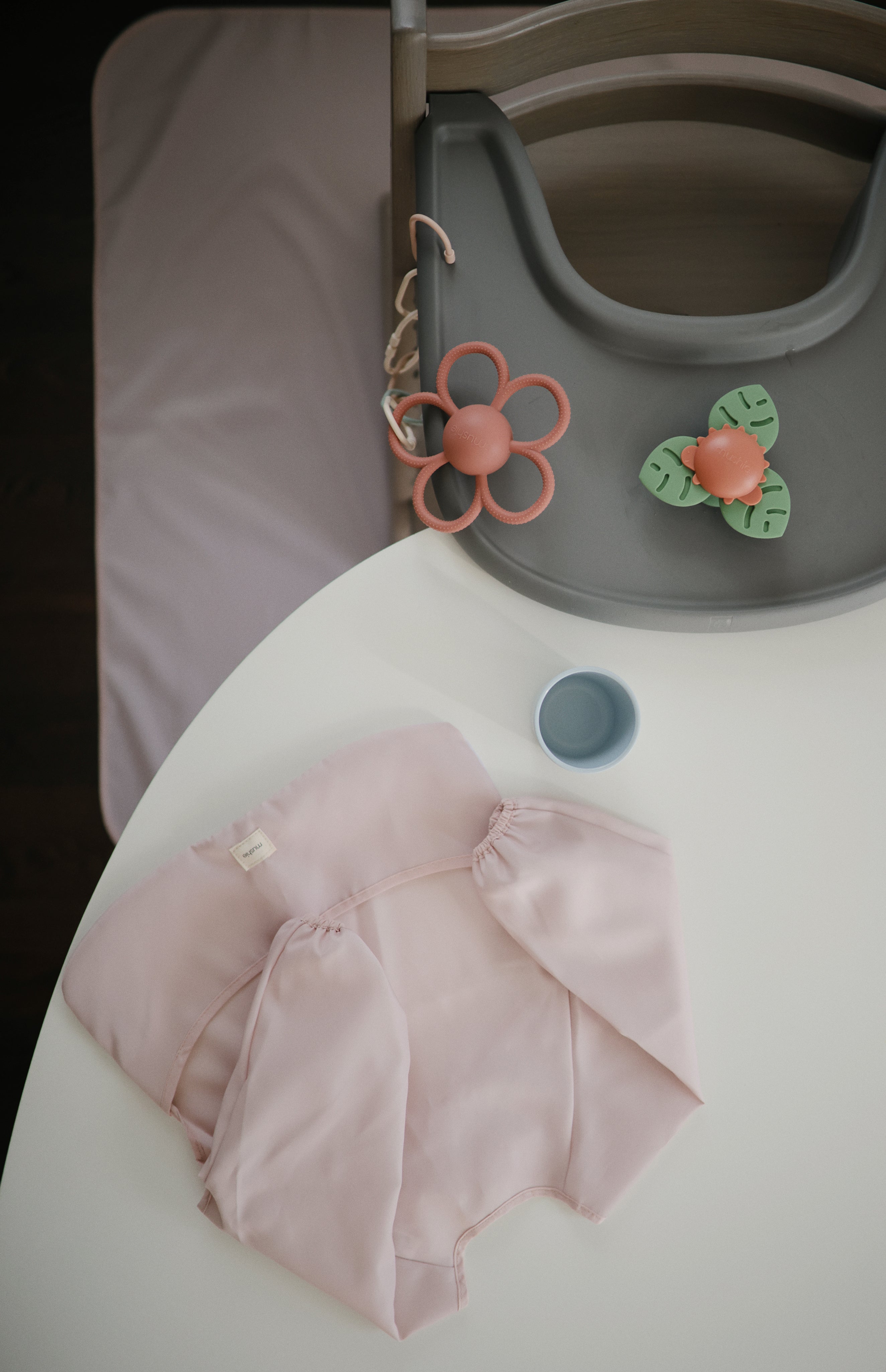 Mushie long sleeve bib in blush laying on a white table styled next to a grey high chair and baby toys