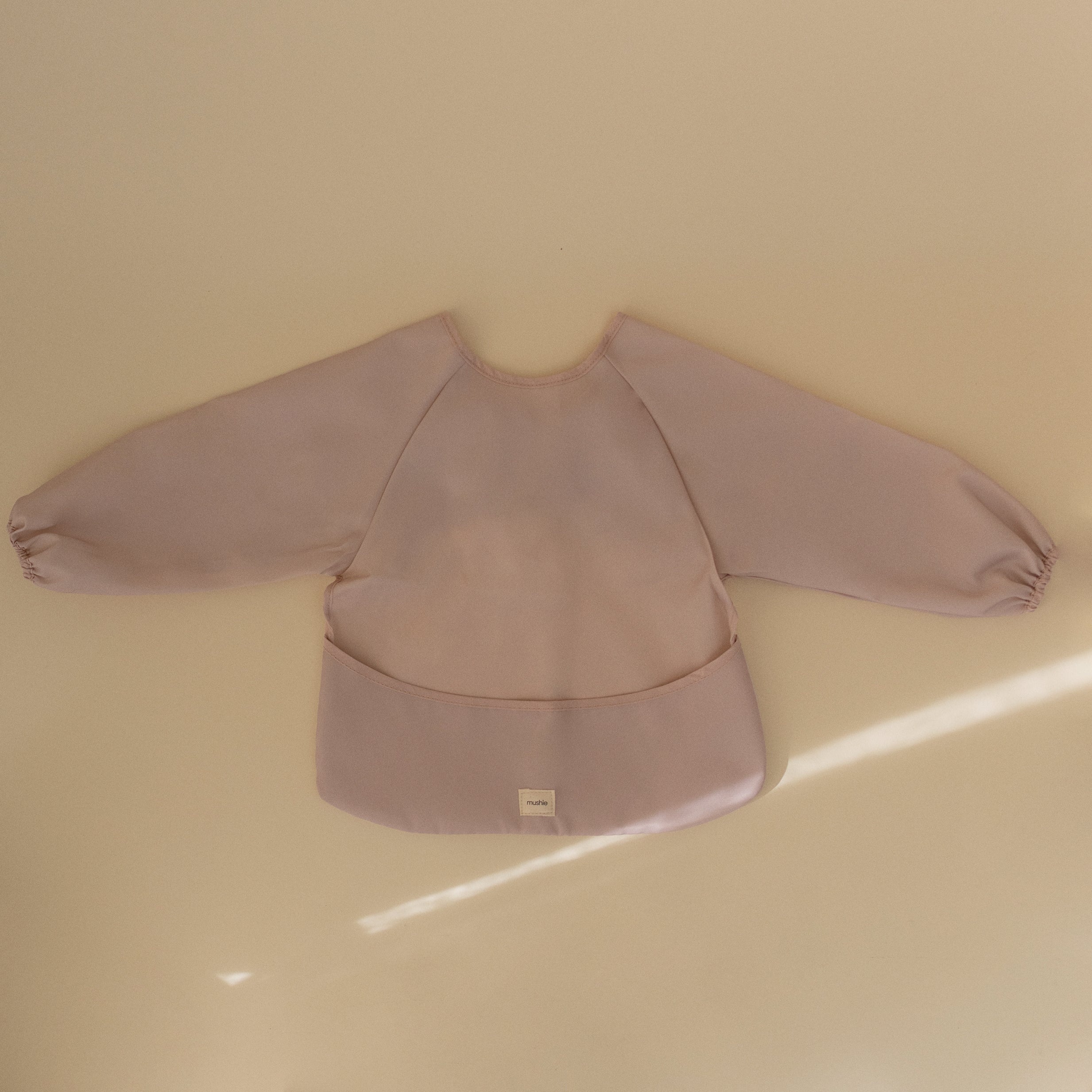 Lifestyle image of a Mushie long sleeve bib in Blush against a beige background