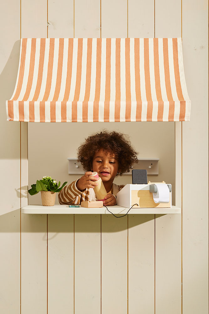 Child playing with a Kid's Concept toy cash register under a striped canopy in a play area