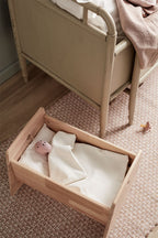 Kids Concept doll crib with a baby doll inside (not included), placed on a textured floor next to a bed