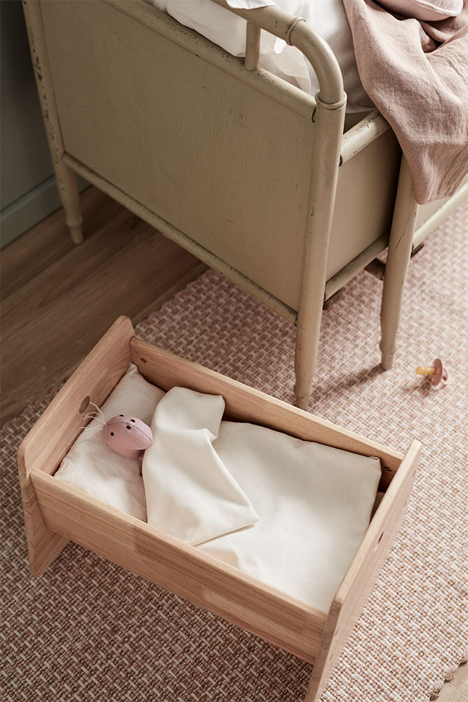 Kids Concept doll crib with a baby doll inside (not included), placed on a textured floor next to a bed