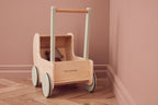Front view of a Kid's Concept doll pram on a wooden floor against a pink wall