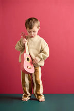 Child holding a Kid's Concept wooden toy guitar on a pink background and a green surface