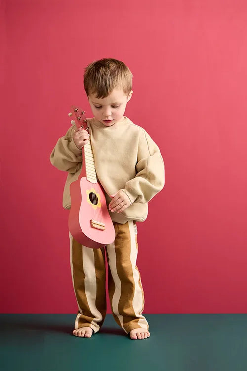 Child holding a Kid's Concept wooden toy guitar on a pink background and a green surface