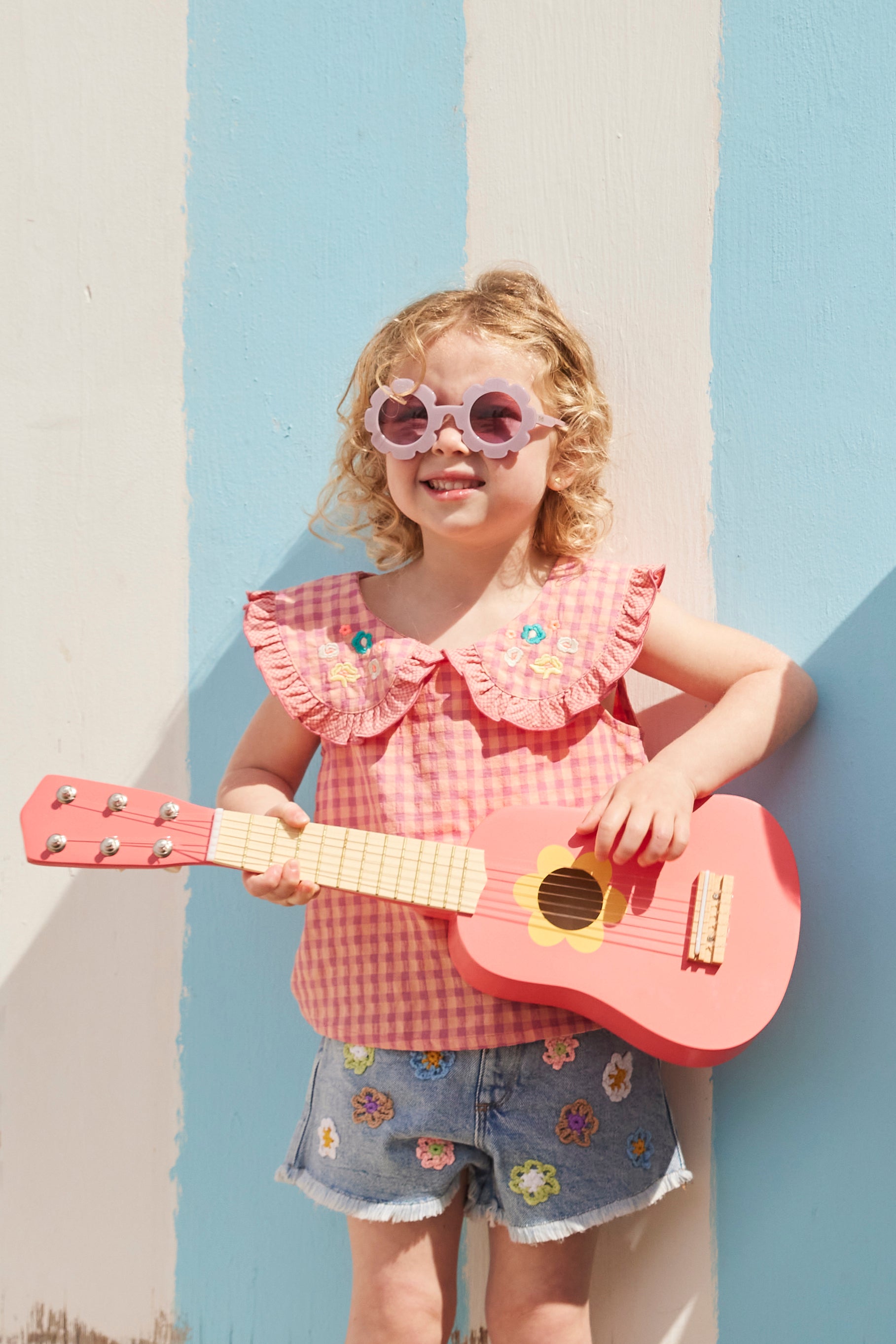 Young girl holding a Kid's Concept wooden toy guitar against a colorful wall
