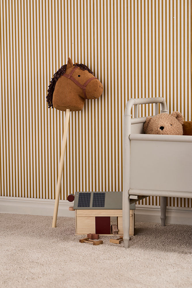 Children's room with a Kid's Concept hobby horse, wooden farm house, and teddy bear against striped wallpaper.