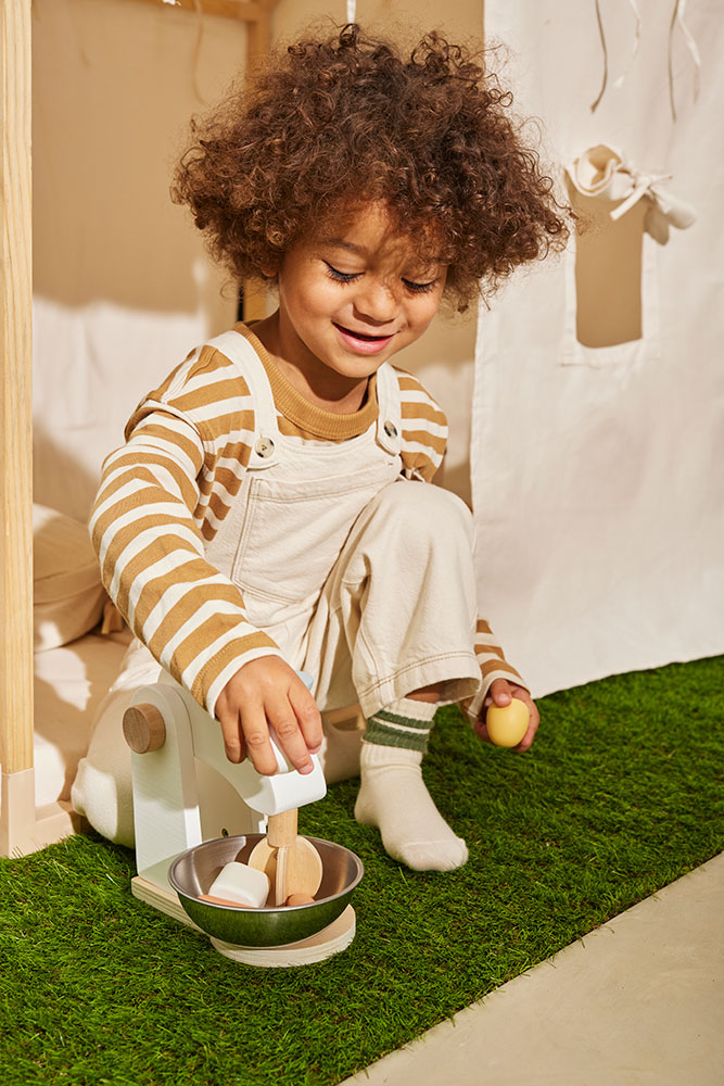 Child playing with the Kid's Concept wooden mixer set on a white background in a play area with green grass-like flooring
