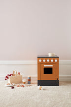 Kid's Concept picnic play set styled next to an orange kid's stove (not included) with black base on a light carpet against a neutral wall