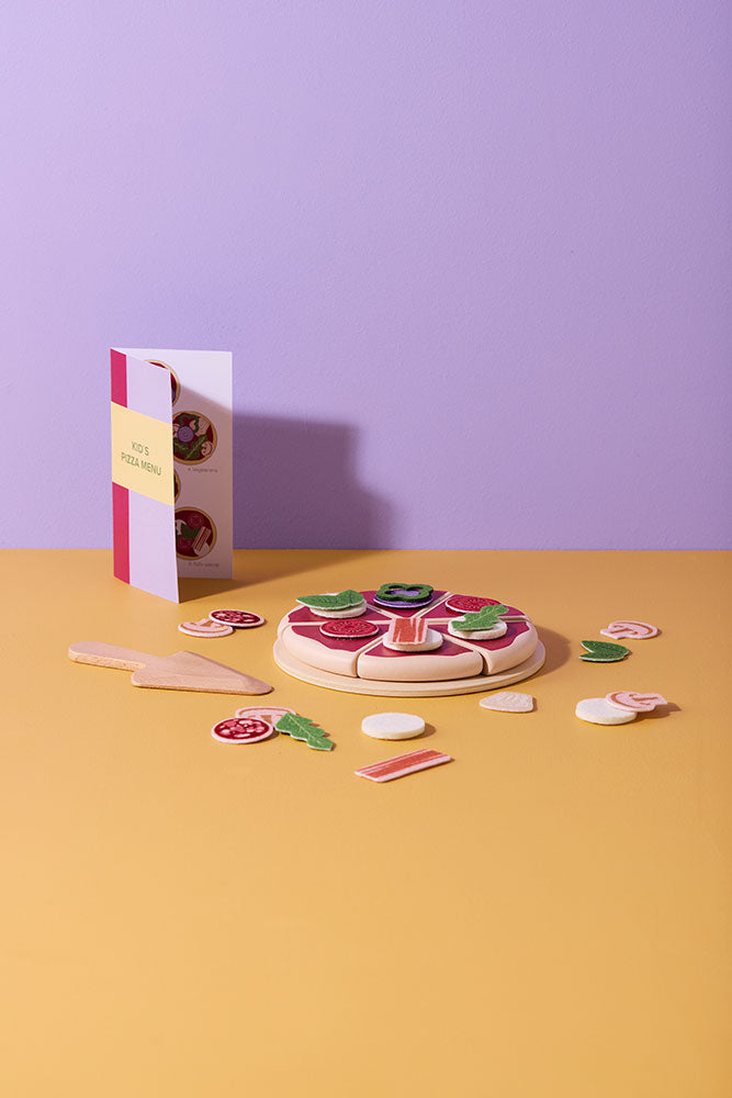 Kids Concept wooden pizza play kit with pieces on a yellow surface against a purple wall
