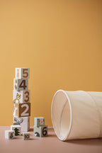 Kid's Concept kids play tunnel in off-white styled next to a stack of wooden blocks with numbers and animal illustrations (not included) on a yellow background