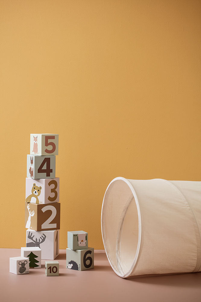 Kid's Concept kids play tunnel in off-white styled next to a stack of wooden blocks with numbers and animal illustrations (not included) on a yellow background