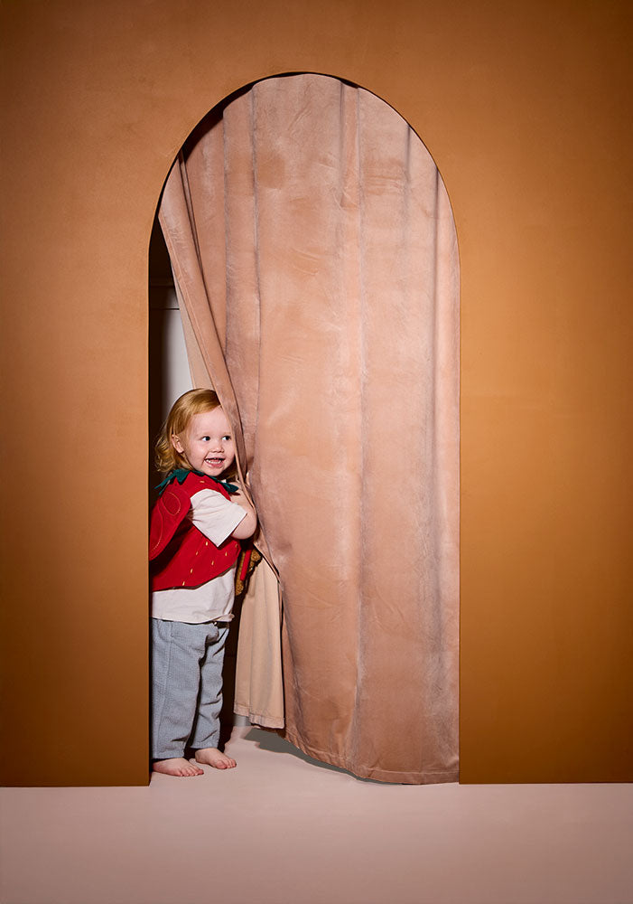Child wearing a Kid's Concept strawberry fairy dress-up costume peeking out from behind a beige curtain in a room with a brown wall