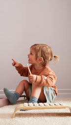 Child playing with Kid's Concept wooden xylophone on a light-colored carpet against a plain wall