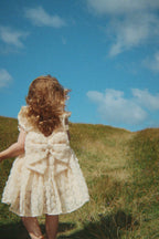 Child wearing the Konges Sløjd Ally dress in Sandshell, while standing on a grassy hill with a blue sky.