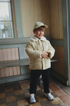 A young boy wearing a Konges Sløjd Alpi Collar jacket in Pale Olive Green, a matching beige baseball cap, black pants, and white sneakers, standing in a vintage indoor setting with wooden walls and tiled floors.