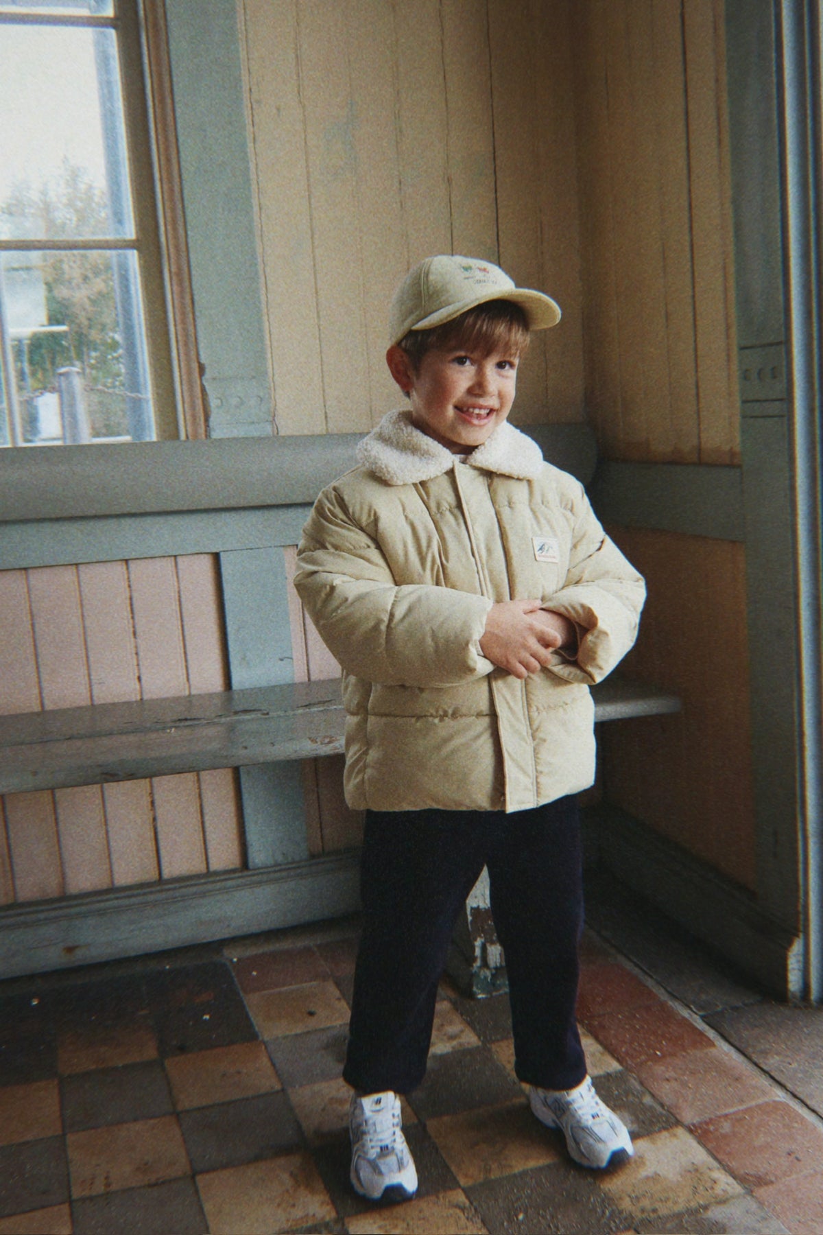 A young boy wearing a Konges Sløjd Alpi Collar jacket in Pale Olive Green, a matching beige baseball cap, black pants, and white sneakers, standing in a vintage indoor setting with wooden walls and tiled floors.