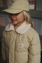 A young girl wearing a Konges Sløjd Alpi Collar jacket in Pale Olive Green and a matching beige baseball cap, standing in front of a rustic wooden wall.