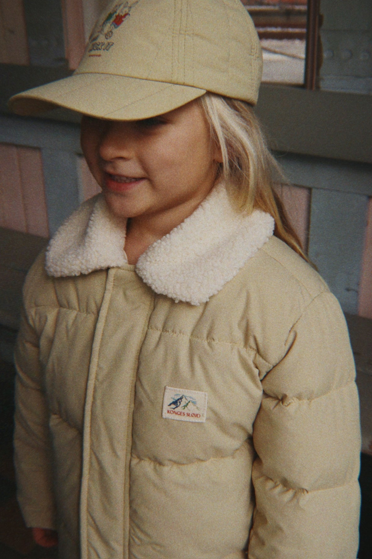 A young girl wearing a Konges Sløjd Alpi Collar jacket in Pale Olive Green and a matching beige baseball cap, standing in front of a rustic wooden wall.
