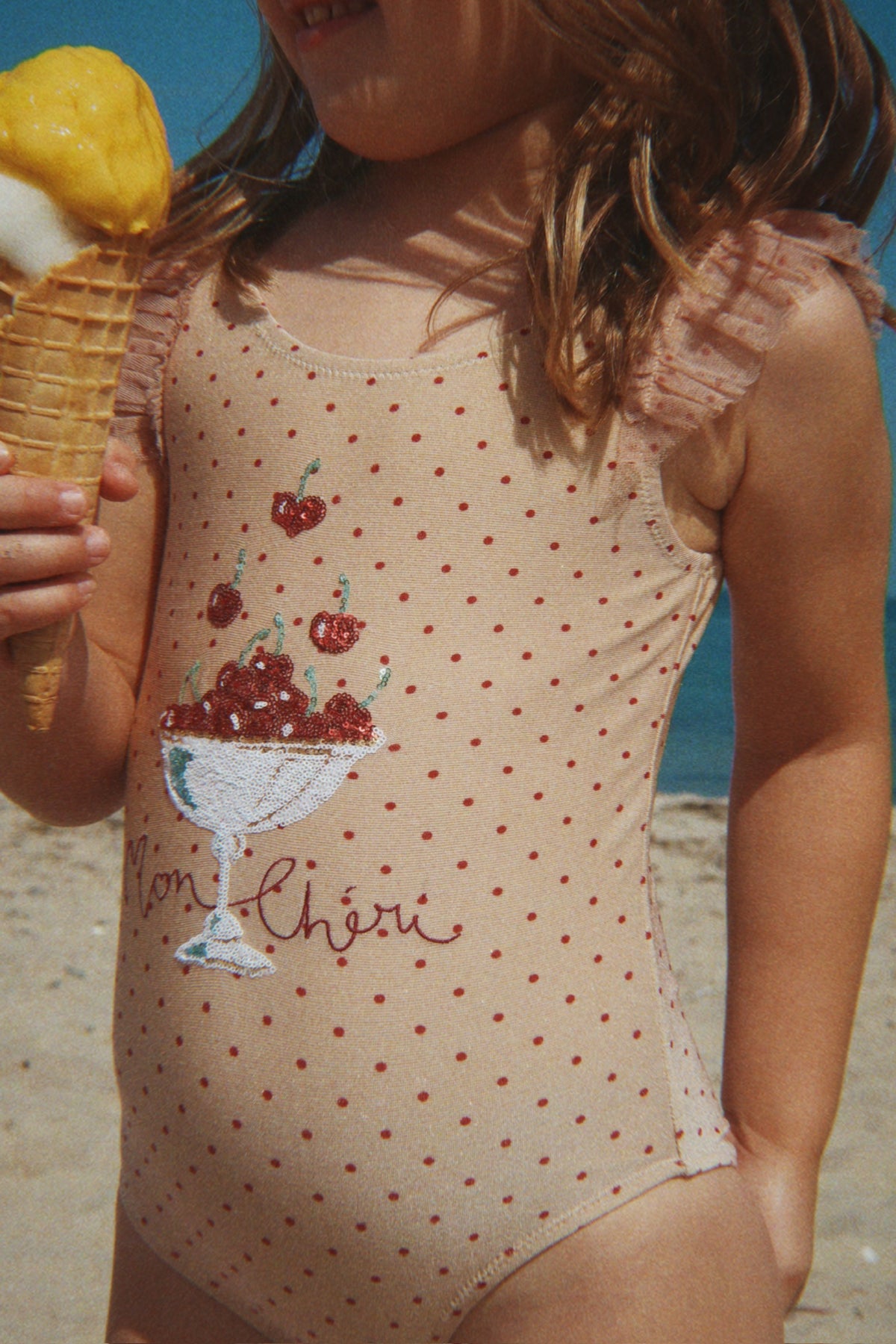 Close-up of a child in a Konges Sløjd Amandine swimsuit in Emboldened not, holding an ice cream cone on a beach.