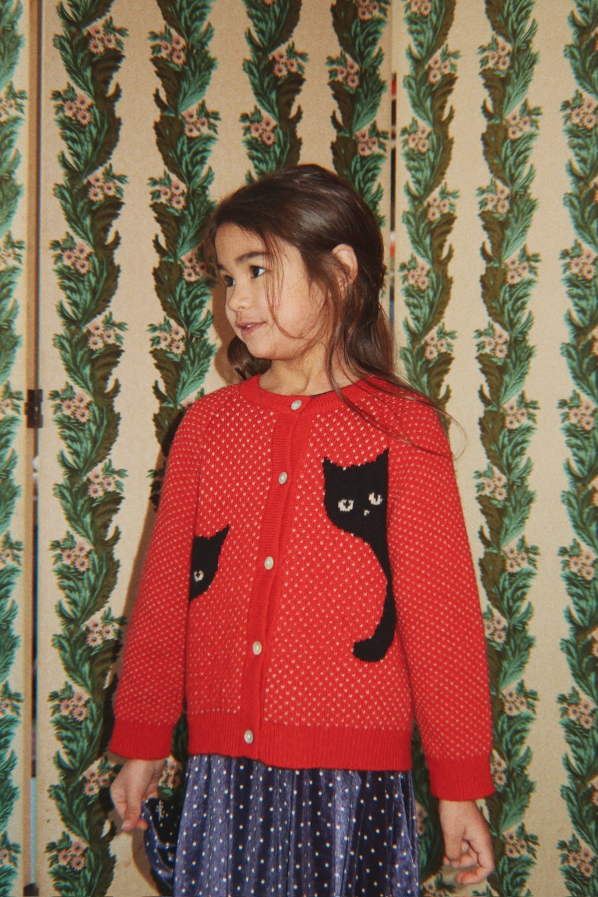 A young girl wearing the Konges Sløjd Belou cardigan in Cat Rouge and a blue polka-dot skirt, standing in front of a vintage green and beige floral wallpapered folding screen.