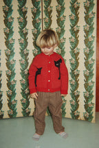 A young child wearing the Konges Sløjd Belou cardigan in Cat Rouge and a brown checkered pants, standing in front of a vintage green and beige floral wallpapered folding screen.