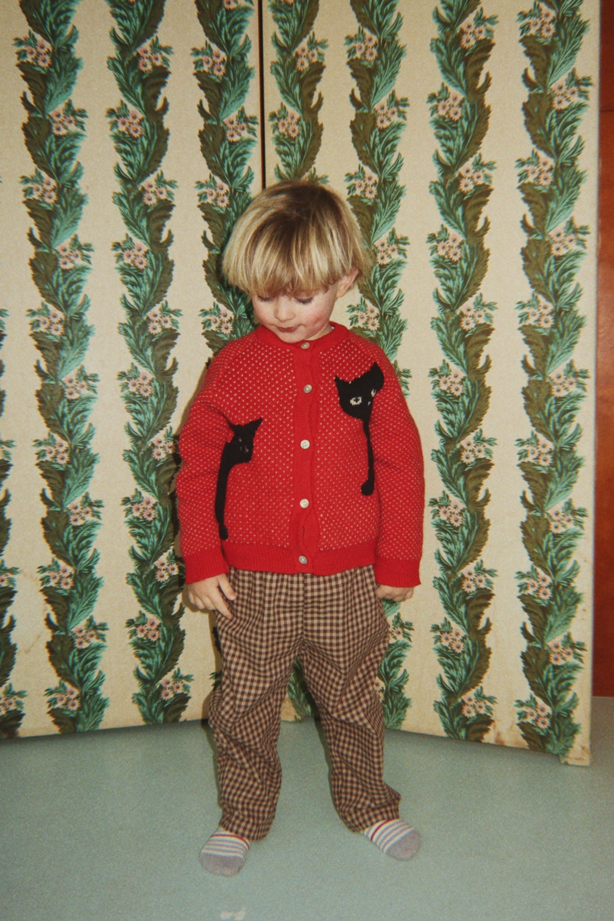 A young child wearing the Konges Sløjd Belou cardigan in Cat Rouge and a brown checkered pants, standing in front of a vintage green and beige floral wallpapered folding screen.