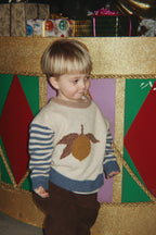 A young child wearing the Konges Sløjd Billi Sweater in Lemon on a white background, standing in front of a festive gold, green, and red display.