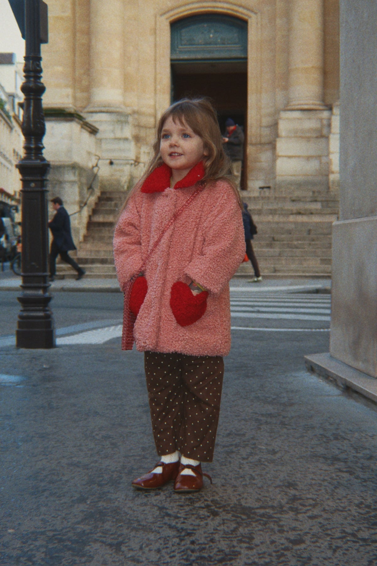 A young girl standing on a city street in front of a stone building, wearing the Konges Sløjd Calin Coat in Mellow Rose, brown patterned pants, and red shoes.