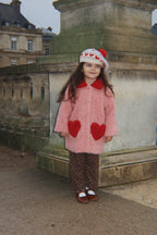 A young girl wearing the Konges Sløjd Calin Coat in Mellow Rose, a white knit hat with red hearts, and brown patterned pants, standing outdoors.