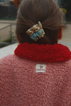 A close-up back view of a person wearing the Konges Sløjd Calin Coat in Mellow Rose. A small white sewn Konges Sløjd label is visible at the center of the collar, and a decorative hair clip with two cats is in the hair.