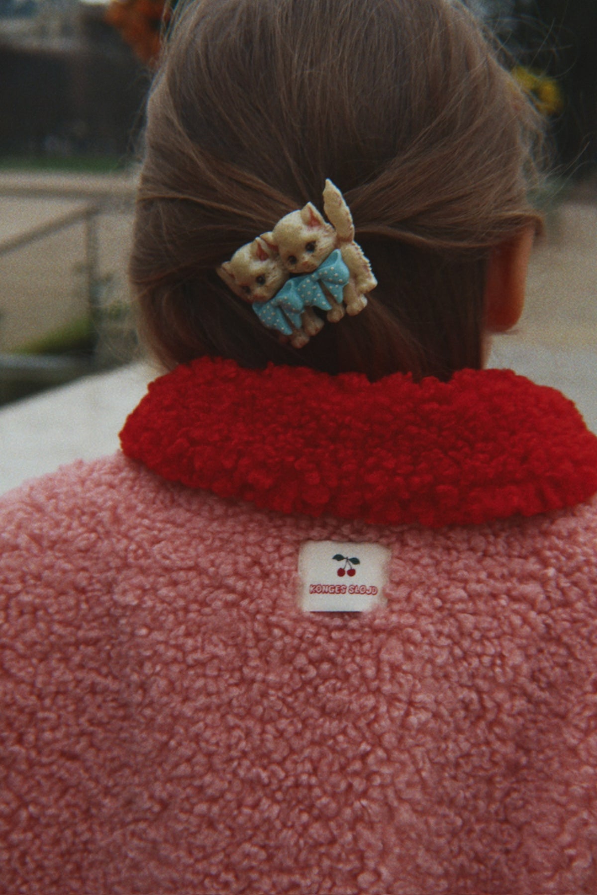 A close-up back view of a person wearing the Konges Sløjd Calin Coat in Mellow Rose. A small white sewn Konges Sløjd label is visible at the center of the collar, and a decorative hair clip with two cats is in the hair.
