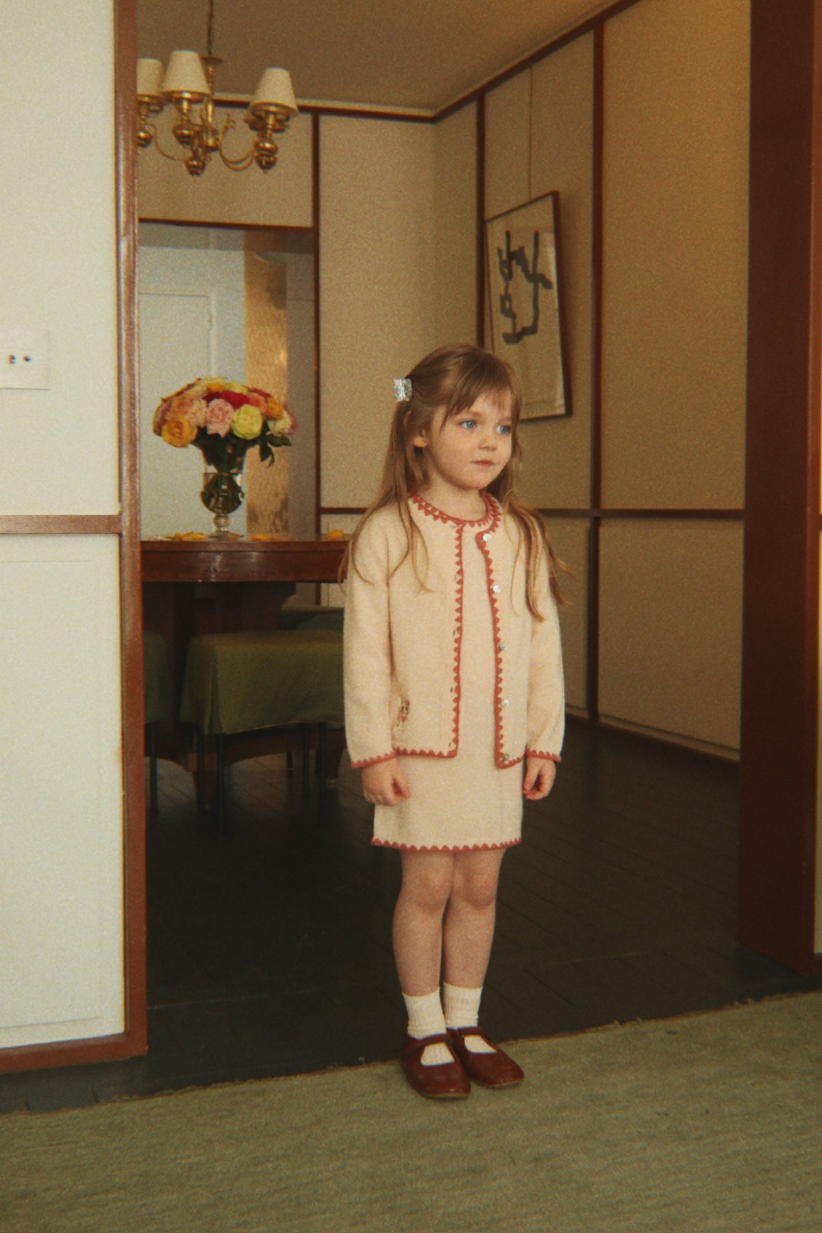 A young girl stands in a doorway wearing the Konges Sløjd Collette Cardigan in Rosey and brown shoes, photographed in an indoor room setting.