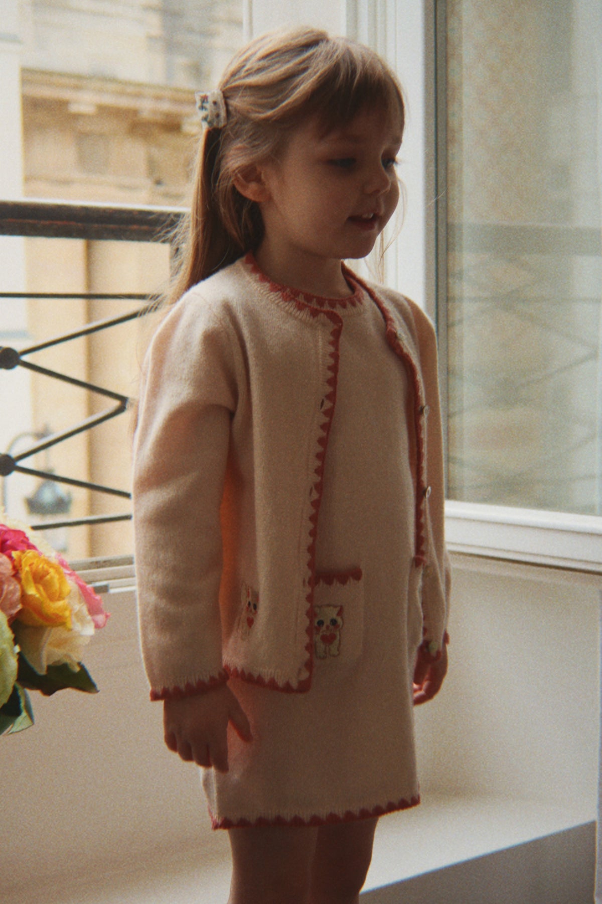 A young girl with light brown hair in a ponytail stands by a window wearing  the Konges Sløjd Collette Cardigan and Collette Dress in Rosey. 