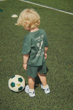 Child in wearing a Konges Sløjd Famo set in Cilantro on a white background standing on a soccer field with a football