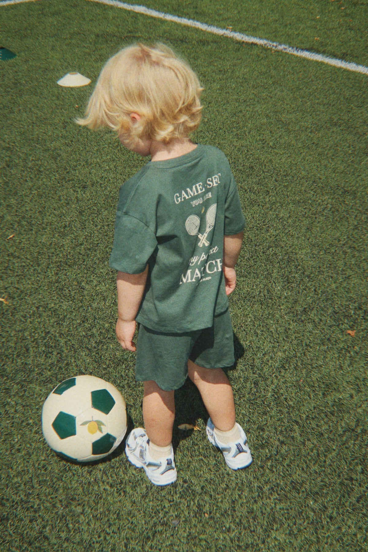 Child in wearing a Konges Sløjd Famo set in Cilantro on a white background standing on a soccer field with a football