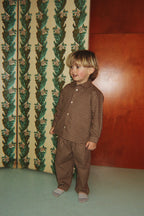 A young child wearing the Konges Sløjd Foret Long-sleeve Shirt and Pants in Chocolate Check, standing in front of a patterned green and cream folding screen and a wooden wall.